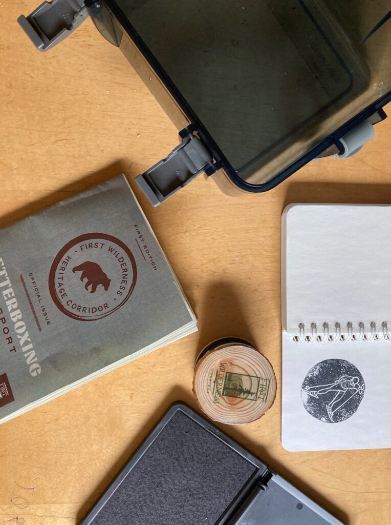 Letterboxing items sit on a wooden tabletop. They include a round wood stamp, a notebook with a stamp in it, a dark gray briefcase, a letterboxing passport, and a black ink pad.
