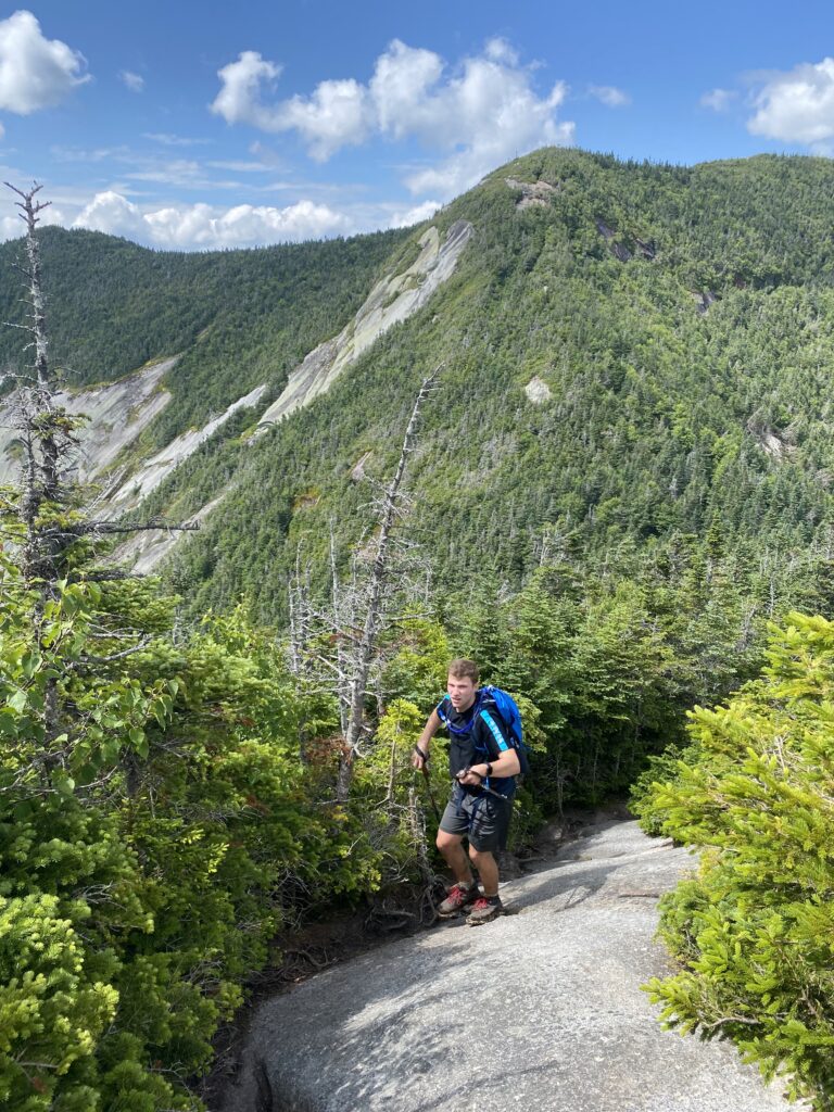 Lush green mountains tower behind a white, blonde-haired man in scaling up a rock. He is wearing black shorts, black t-shirt, and black hiking boots.