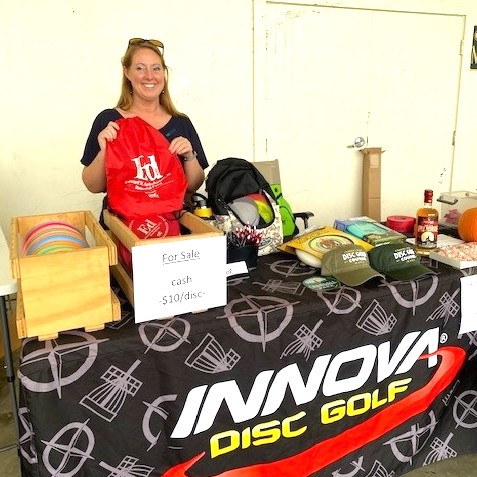 A table is set up with a banner that reads "Innova Disc Golf." A blonde woman stands behind it holding up a red bag that has the Bartholomew logo on it. Various merch is on the table.