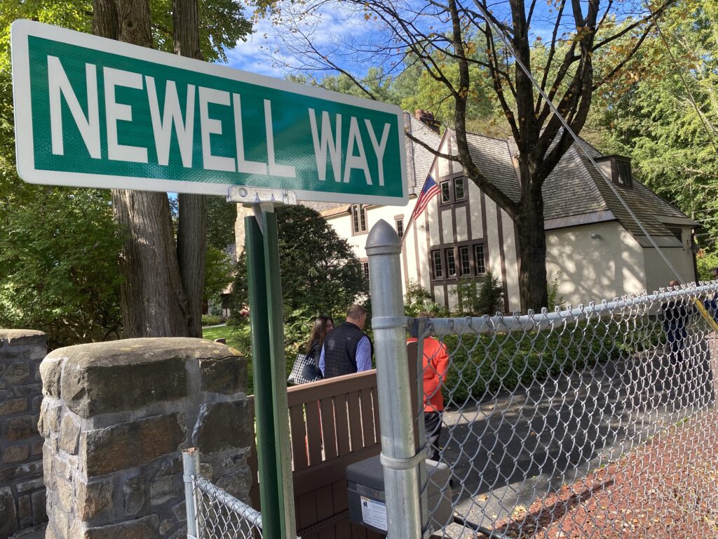 Close up of the sign. White text reads, NEWELL WAY in caps against a green backround. You can see part of the fenced path off to the right and the Newell home in the background.
