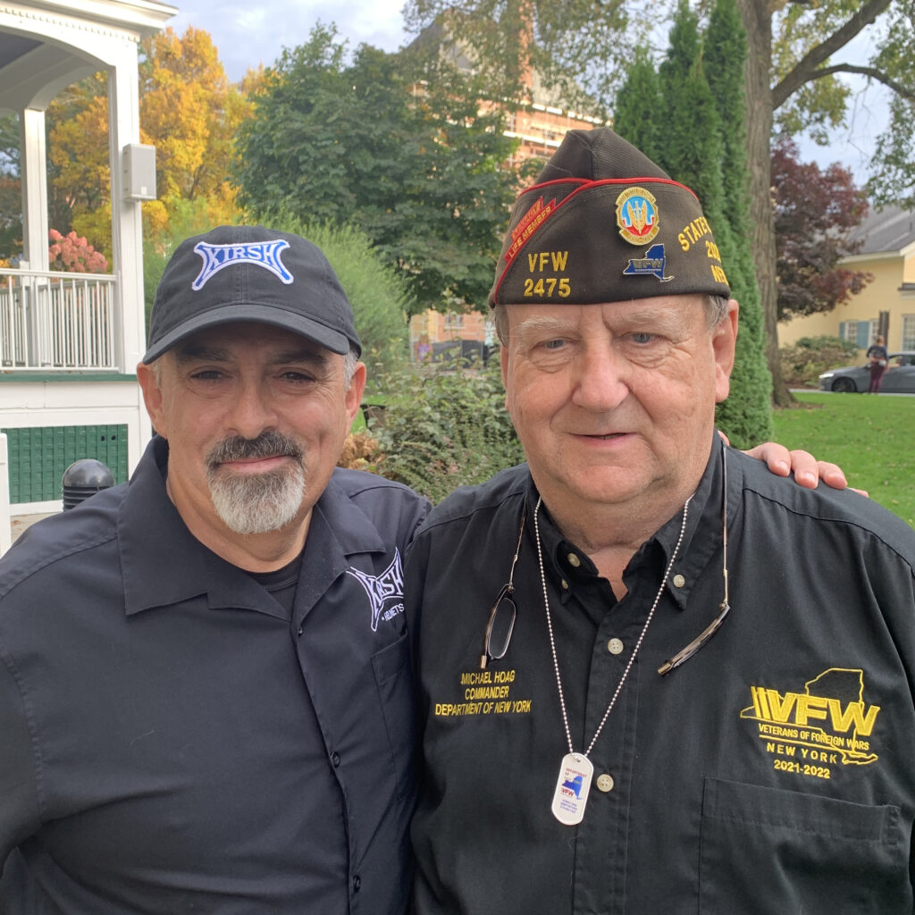 A white middle aged man in a KIRSH shirt and hat has his arm around an older white man in VFW hat and shirt.