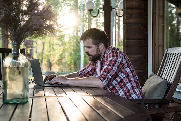 Focused bearded man works with a laptop on the outdoor terrace in his yard, against the background of trees, on a summer evening.