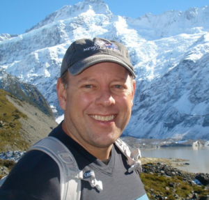 Jim smiles into the camera. He wears a baseball cap and stands outside in front of snow capped mountains.