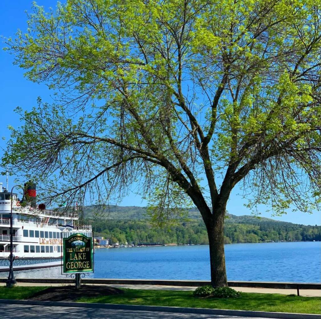 A cruise boat sits at the dock with the blue lake water around it. On the right is a pretty tree and a green sign that says Village of Lake George